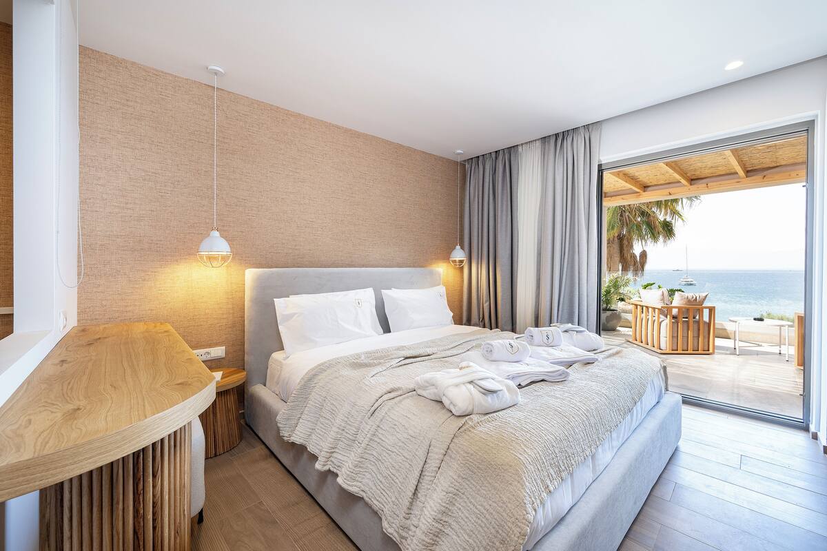A modern hotel room with a large bed, a wooden side table, and a view of the sea through a sliding glass door.