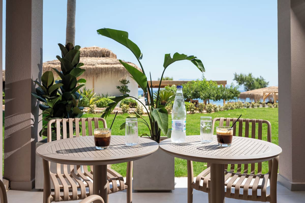 Two round tables with chairs, a bottle of water, and two glasses of iced coffee on a patio with a view of a thatched-roof building and a grassy area.