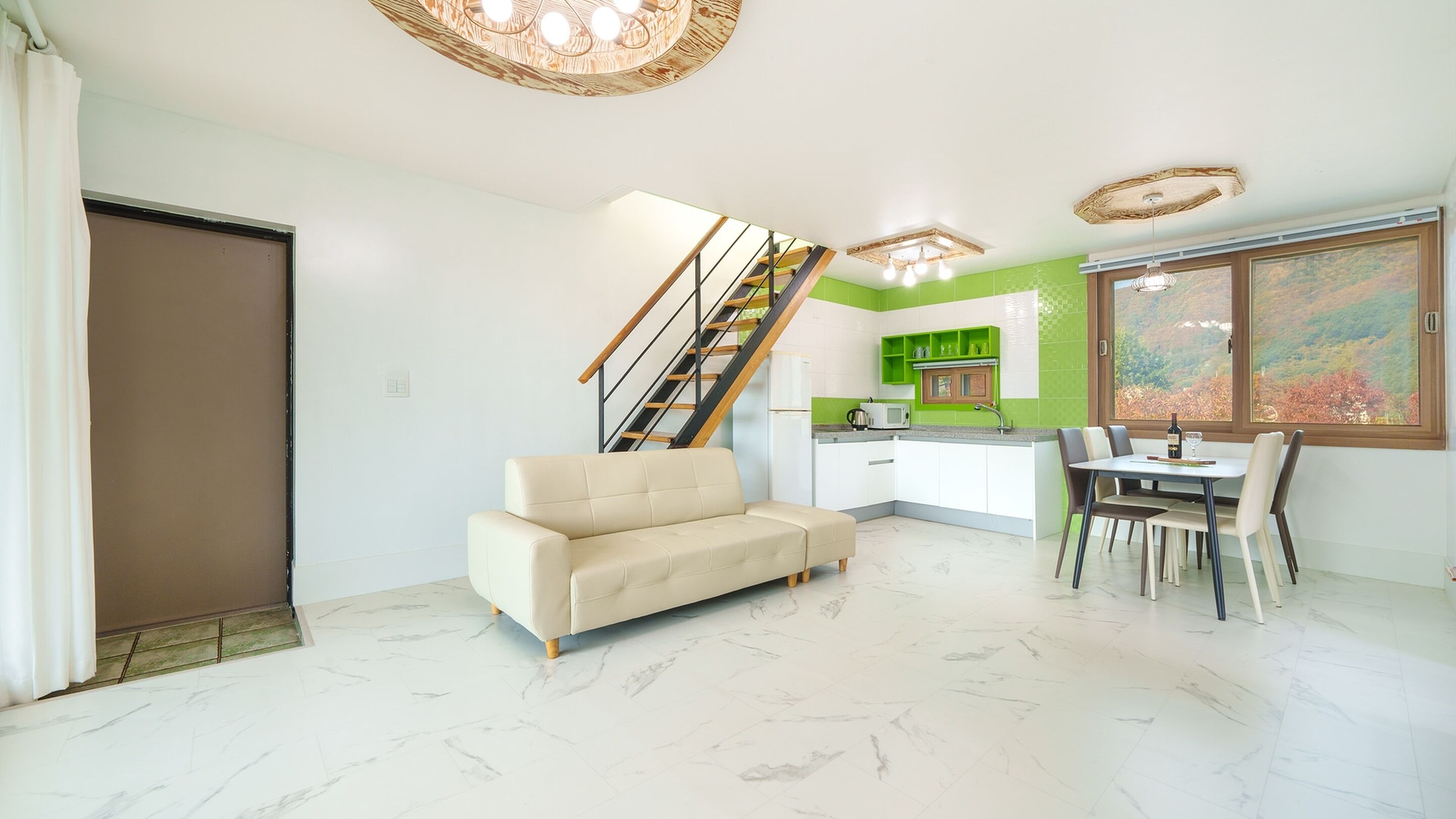 A modern living room with a sofa, dining table, and chairs, a kitchen with green accents, and a staircase.
