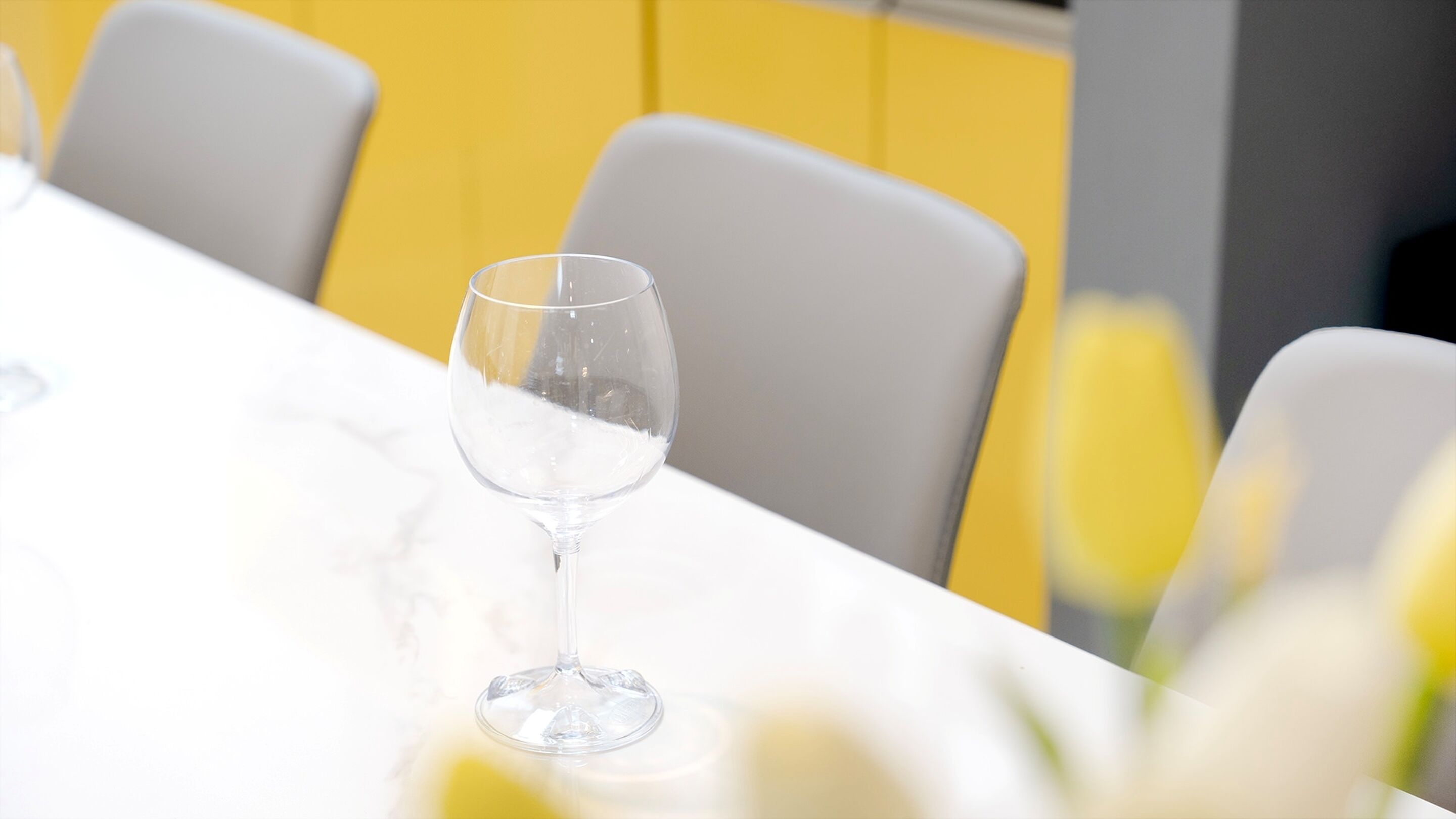 A table with a clear glass and a white chair in a room with yellow walls.