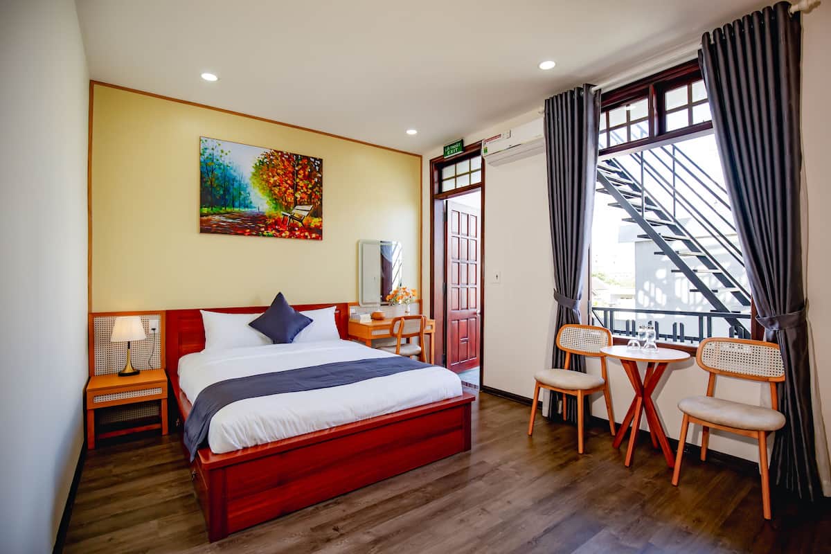 Une chambre d’hôtel avec un grand lit, un bureau, deux chaises, un tableau et une vue sur l’escalier.