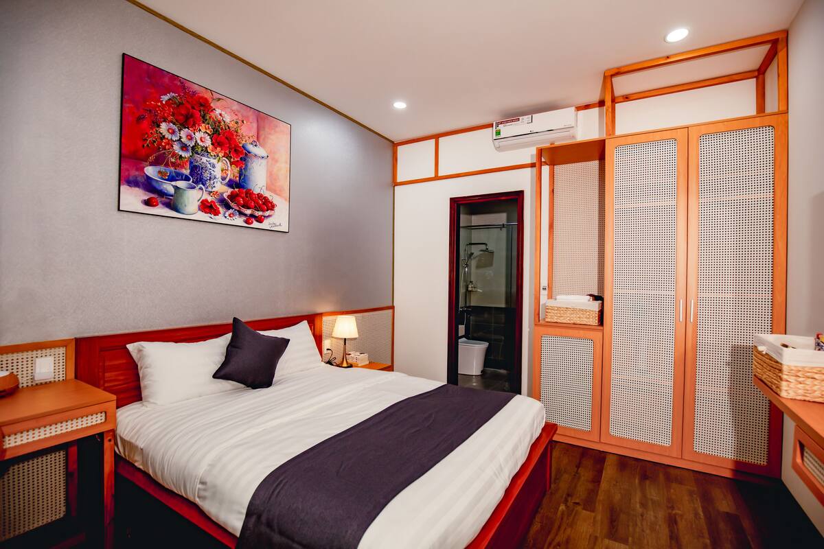 Une chambre d’hôtel comprenant un lit, une table de chevet, une lampe, un tableau, une armoire et une salle de bain visible à travers une porte en verre.