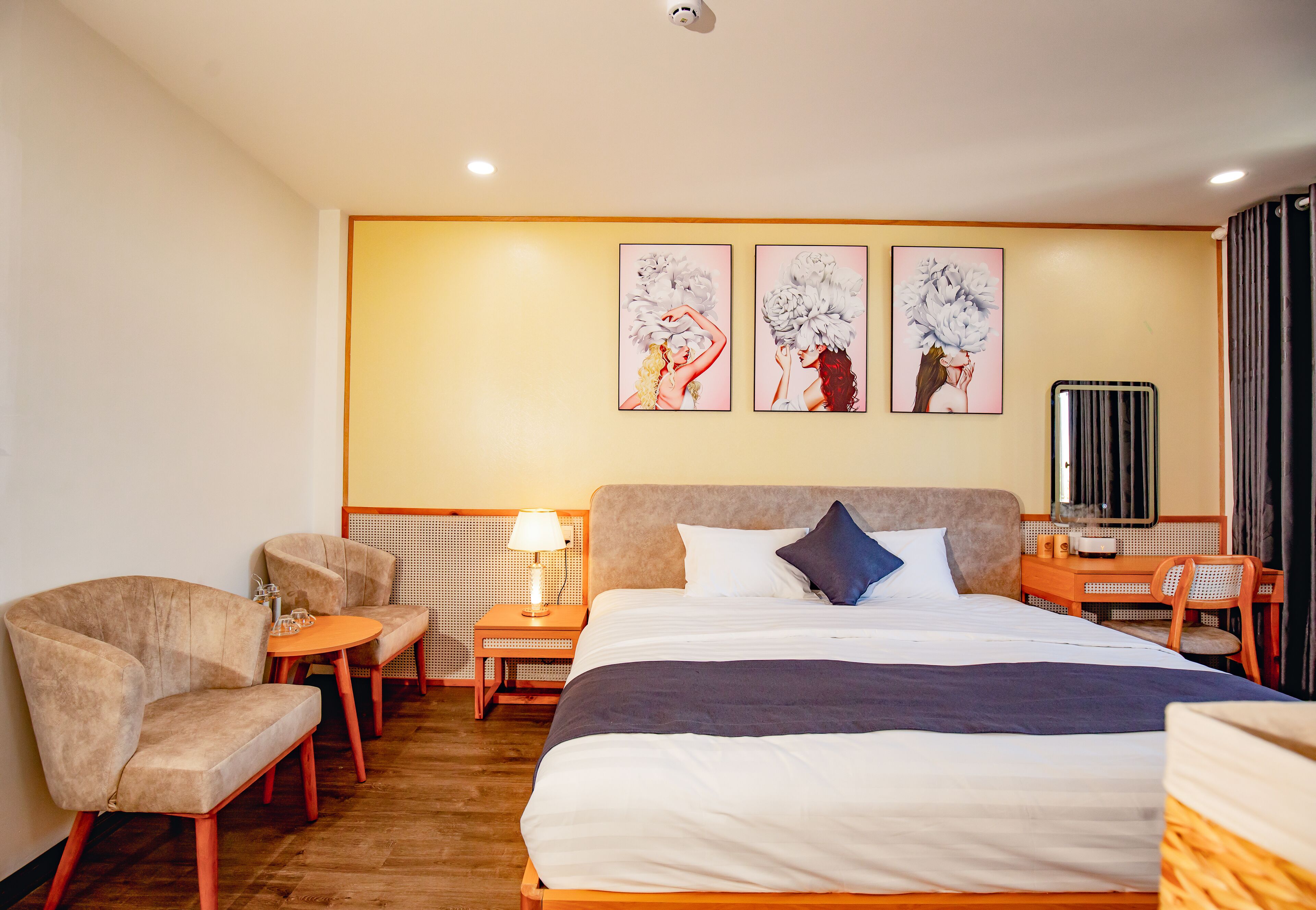 Une chambre d’hôtel avec un grand lit, deux fauteuils, une petite table et un bureau avec une chaise. Trois œuvres d’art encadrées sont accrochées au mur au-dessus du lit.