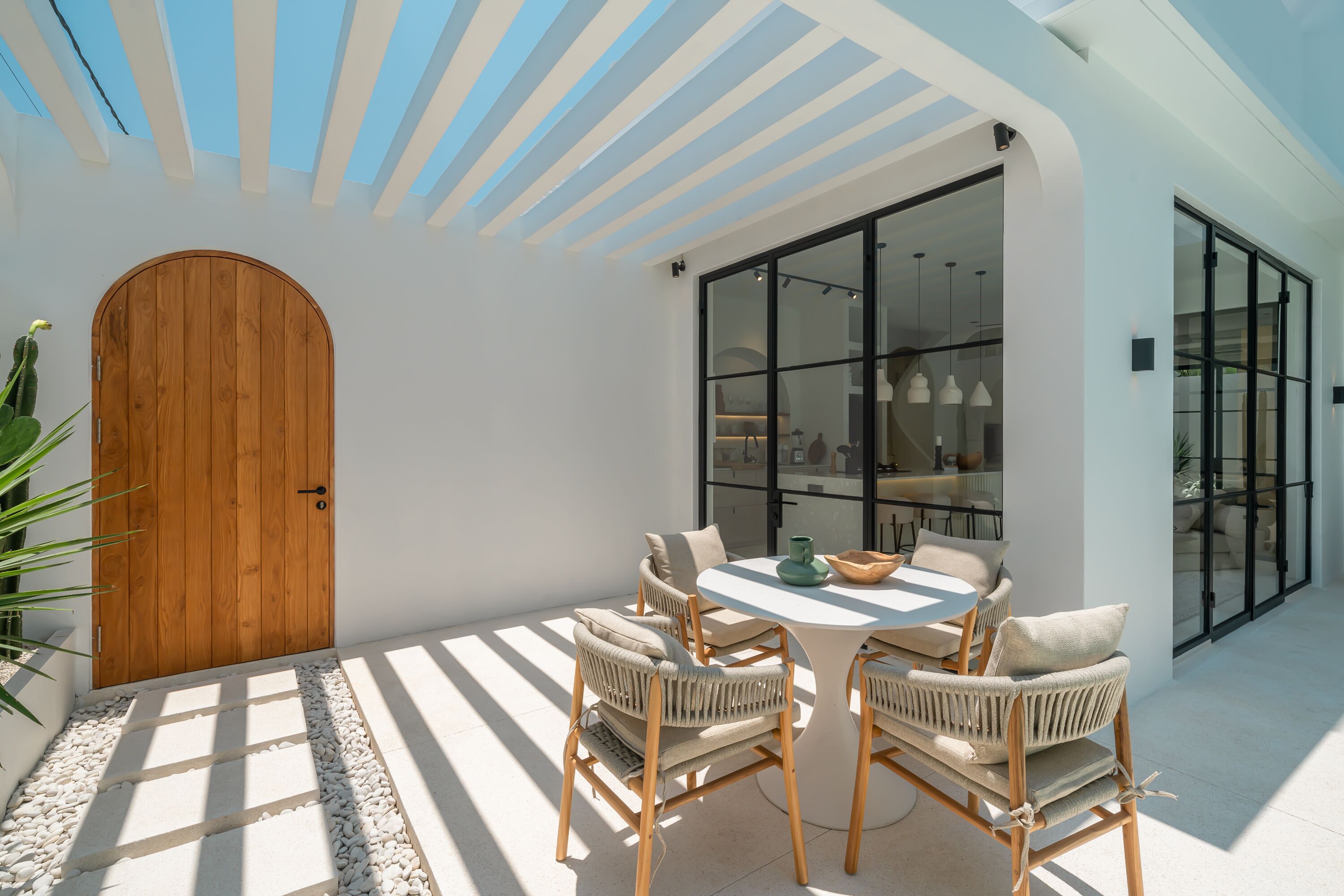 A sunlit patio with a wooden door, wicker chairs, a round table, and a glass door leading to a room.