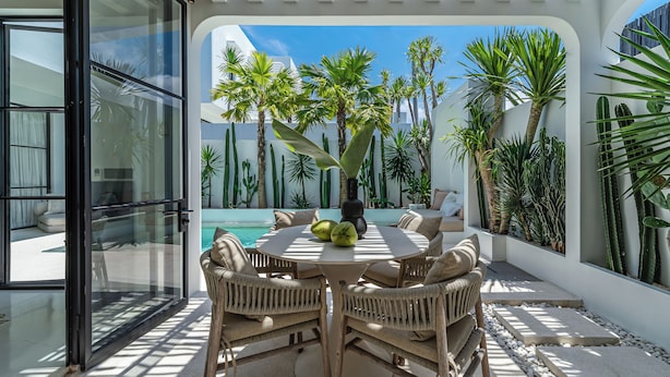 A modern outdoor patio with a round table and wicker chairs, surrounded by various tropical plants and a clear blue sky. - Sanctum house (Kerobokan)