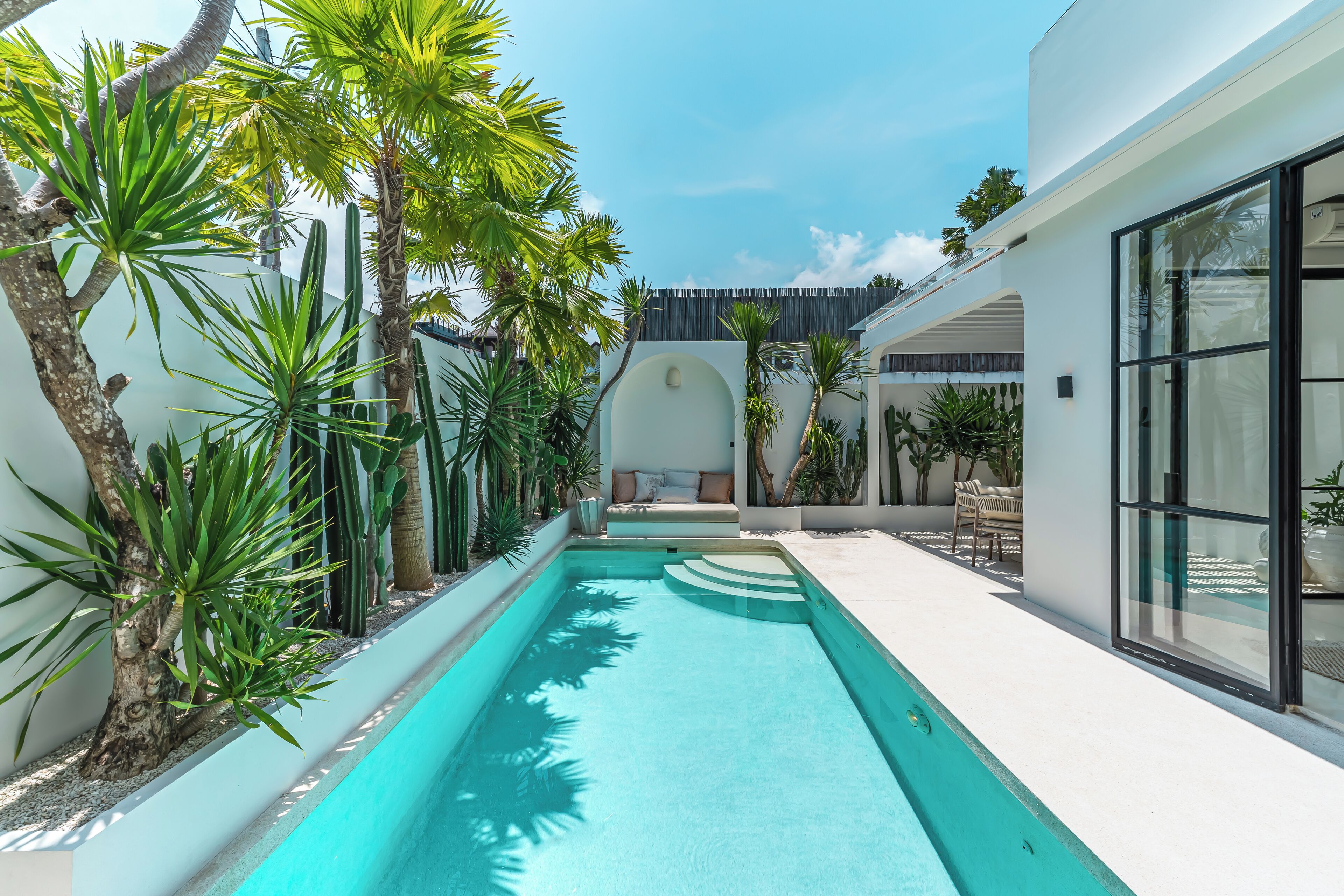 A modern pool area with a clear blue pool, surrounded by tropical plants and a white building with large windows.