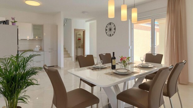 A modern dining area with a white table, six brown chairs, and pendant lights. A clock is on the wall, and a plant is in the foreground. - Amazing home in Marcana (Marcana)