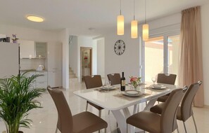 Un espace repas moderne avec une table blanche, six chaises marron et des luminaires suspendus. Une horloge est accrochée au mur, et une plante se trouve au premier plan.