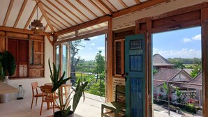 A wooden-framed room with a view of a green landscape, a table with chairs, and a potted plant. - Spaces Bali (Munggu)