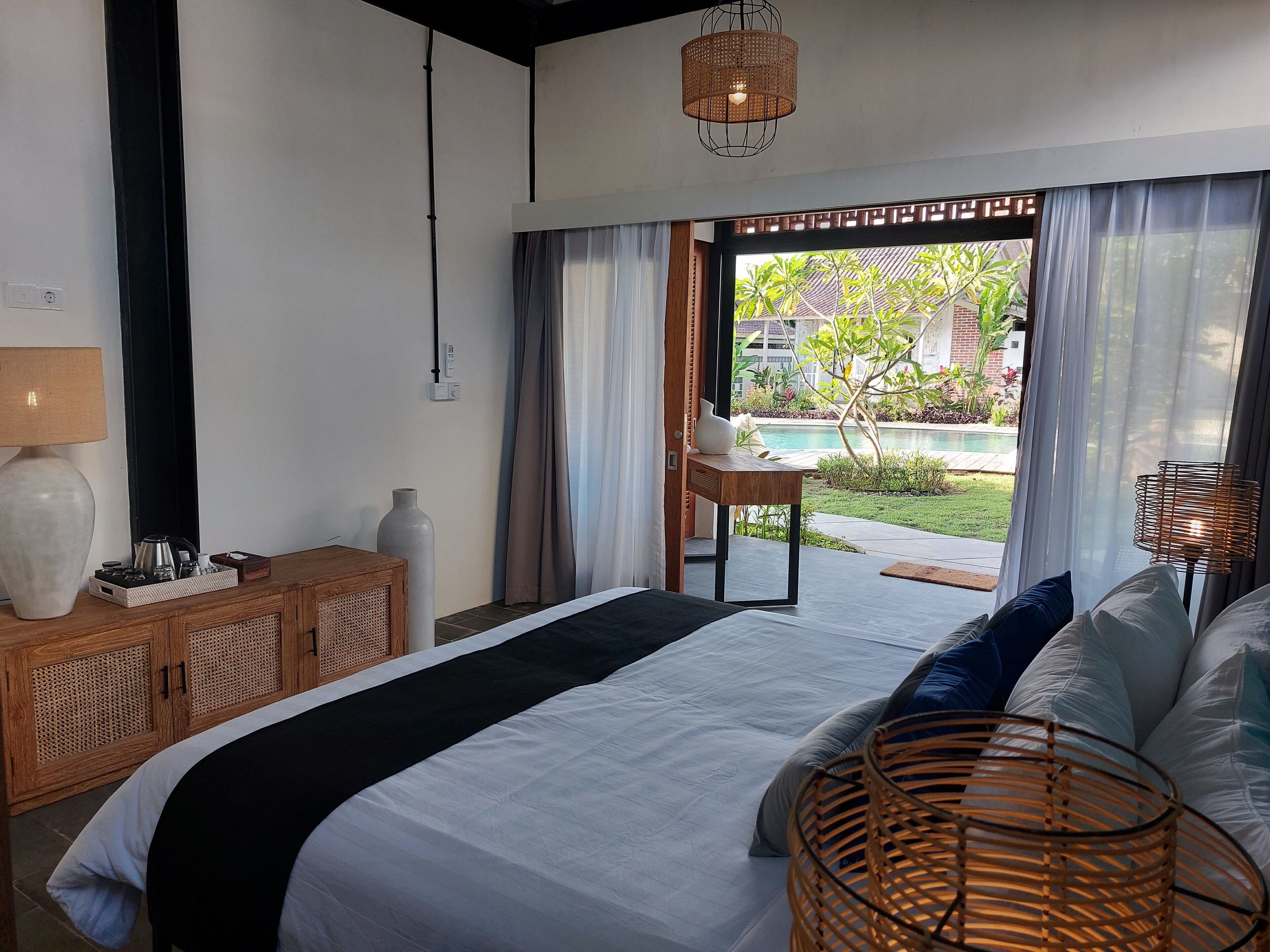 A bedroom with a large bed, a wooden nightstand, a lamp, and a view of a pool and garden.
