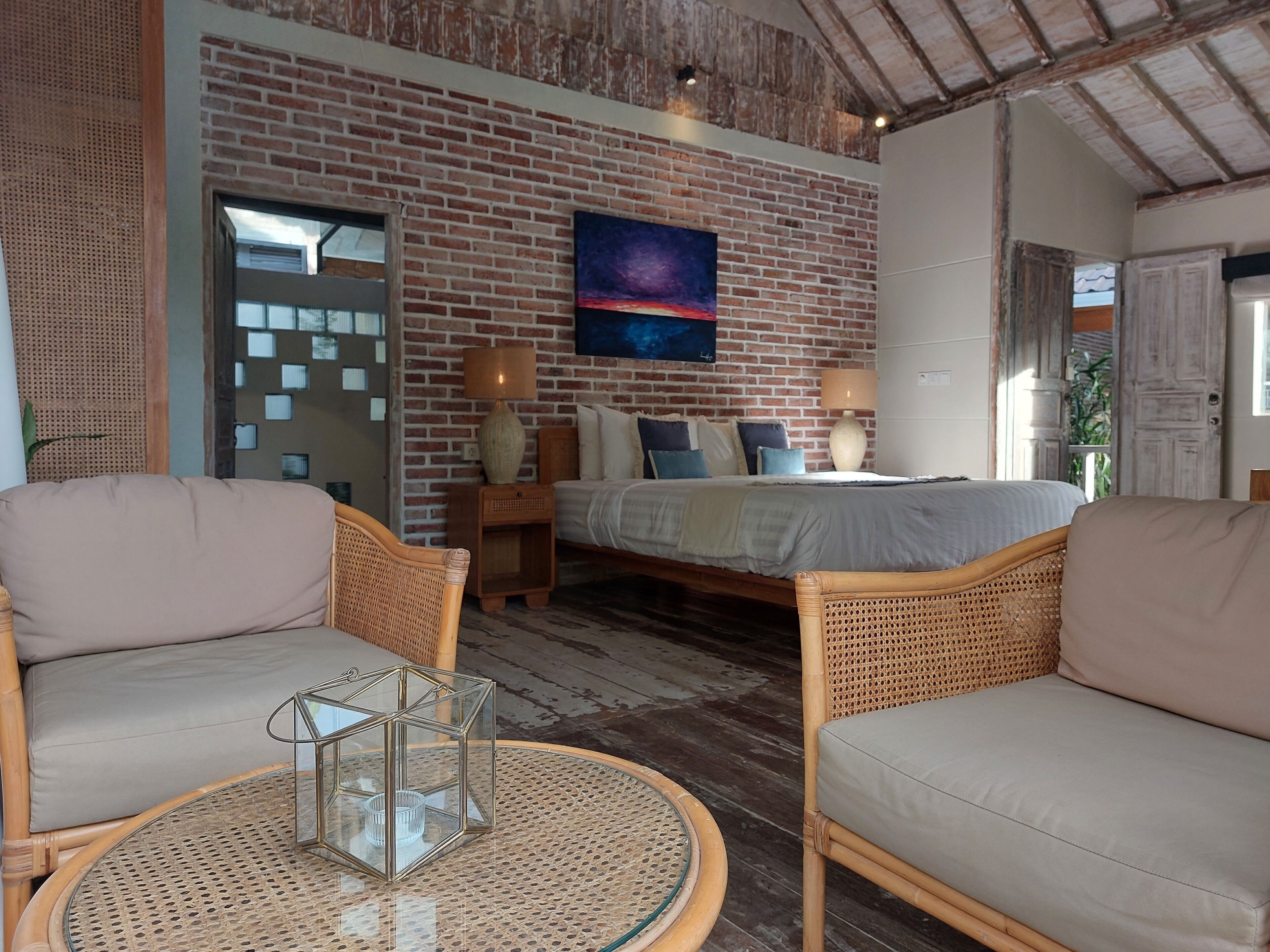 Una habitación acogedora con pared de ladrillo, una cama, dos sillas de mimbre, una mesa redonda con superficie de vidrio y un cuadro sobre la cama.
