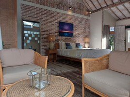 Una habitación acogedora con pared de ladrillo, una cama, dos sillas de mimbre, una mesa redonda con superficie de vidrio y un cuadro sobre la cama.