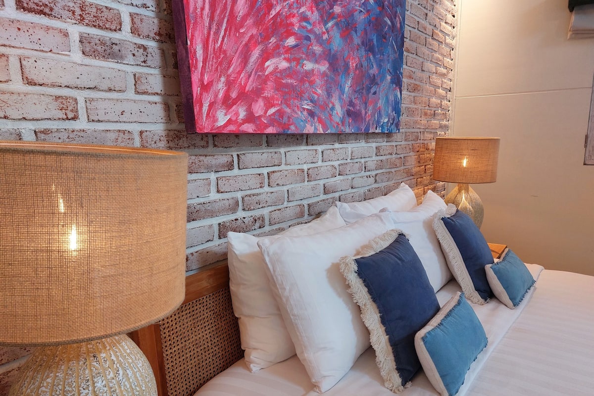A bedroom with a brick wall, a large abstract painting, a bed with white and blue pillows, and a textured lamp.