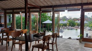 An indoor dining area with wooden tables and chairs, large windows, and a view of an outdoor pool area. - Spaces Bali (Munggu)