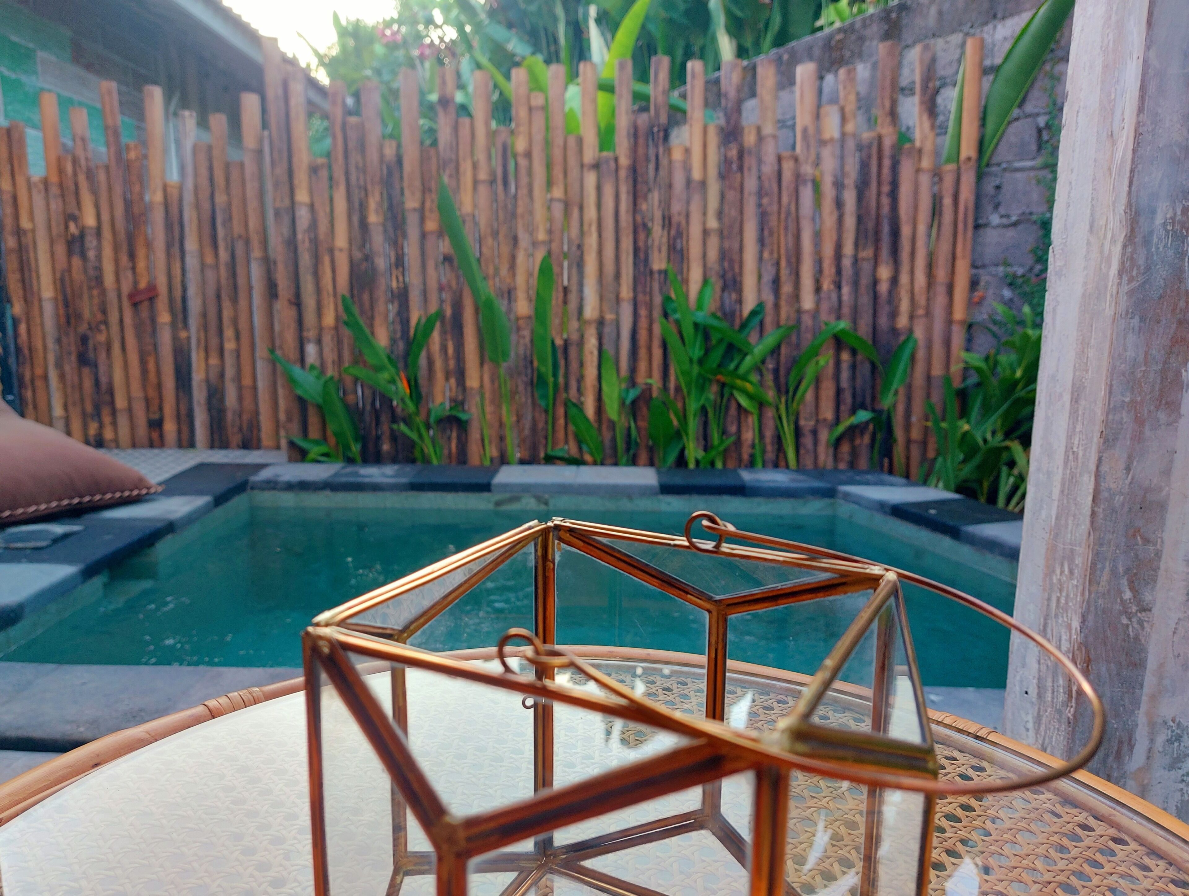 A glass-top table with a copper wireframe centerpiece, a wooden fence, and a pool in the background.