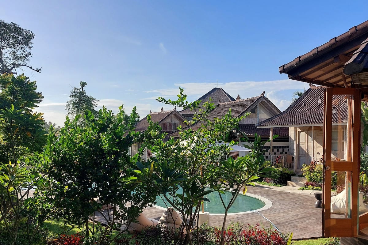 A pool area with a wooden deck, surrounded by lush greenery and a clear blue sky.