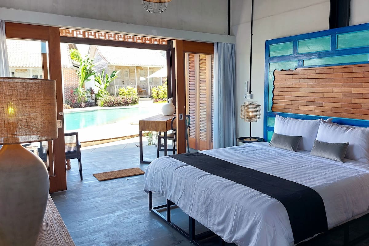 A bedroom with a large bed, a view of a swimming pool, and a balcony with a chair.