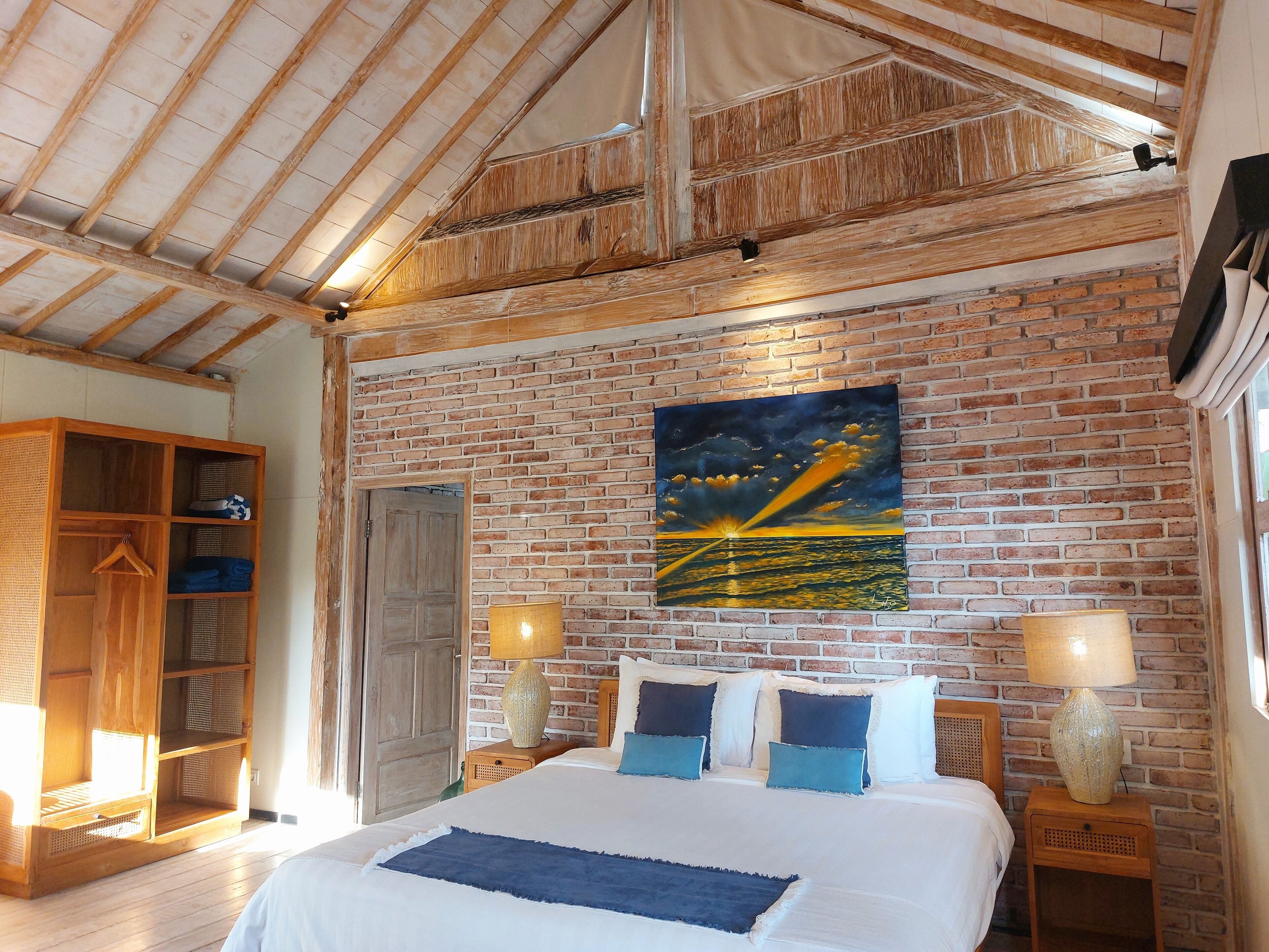 A bedroom with a large bed, a wooden wardrobe, a brick wall, and a painting of a sunset over the ocean.