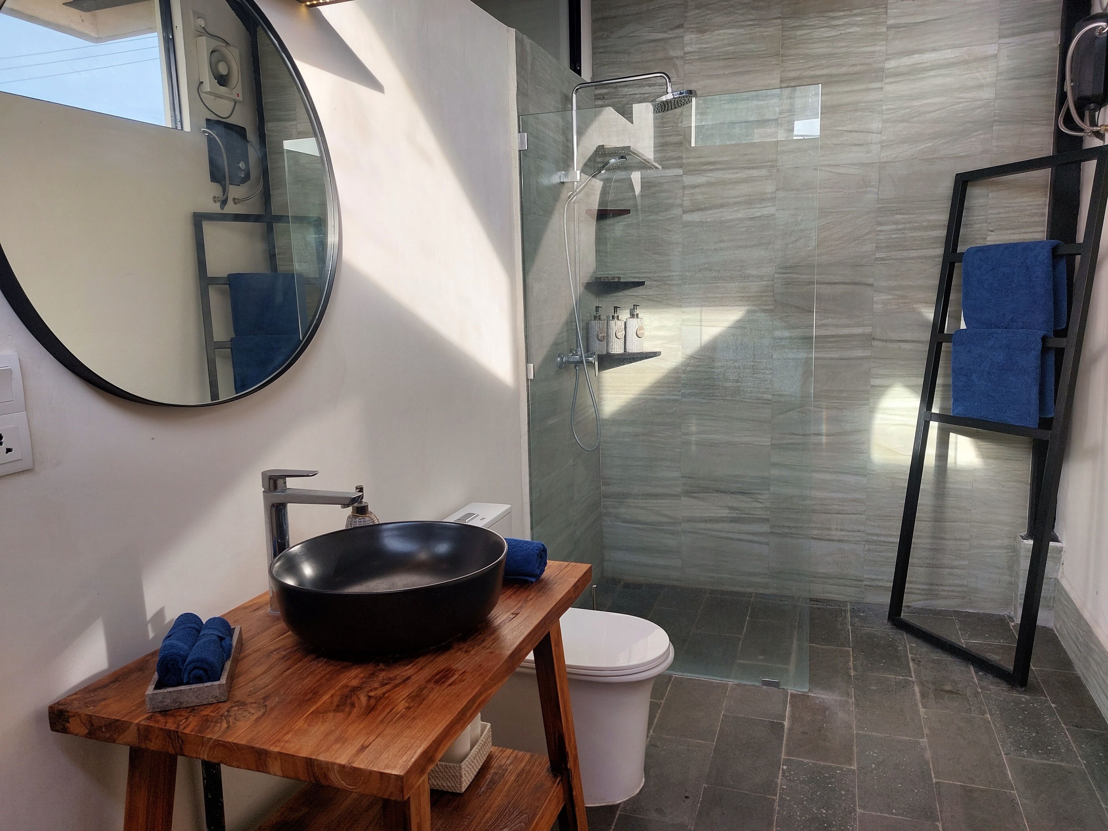 Une salle de bain avec un miroir rond, un meuble-lavabo en bois, un lavabo noir, des toilettes blanches, une cabine de douche en verre et un porte-serviettes bleu.