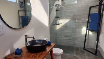 Une salle de bain avec un miroir rond, un meuble-lavabo en bois, un lavabo noir, des toilettes blanches, une cabine de douche en verre et un porte-serviettes bleu.