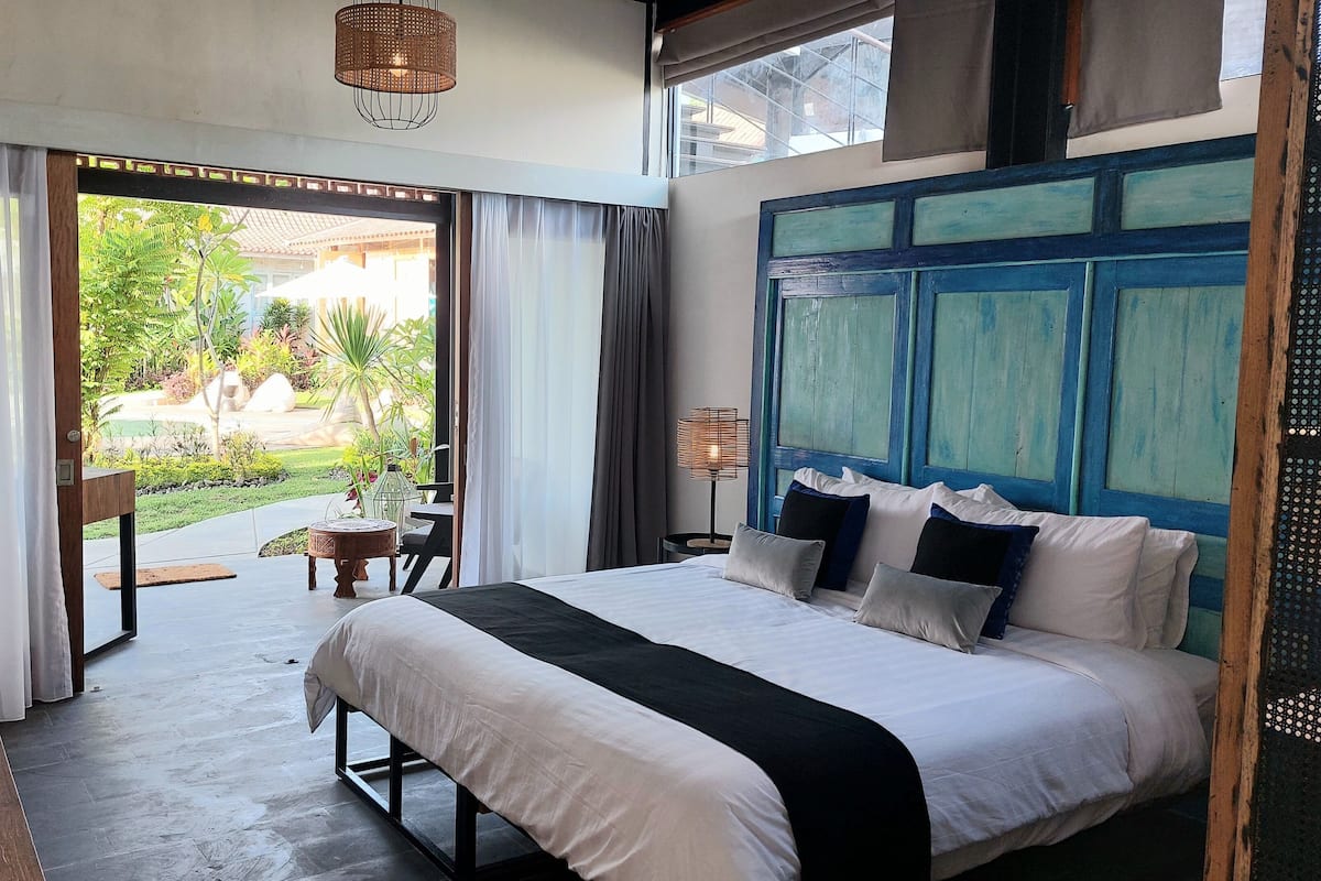 A hotel room with a bed, a lamp, a view of the garden, and a window with curtains.