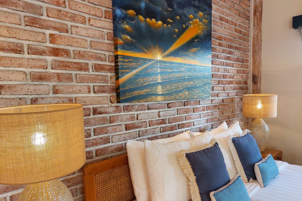 A hotel room with a brick wall, a painting of a sunset over the ocean, a bed with white and blue pillows, and two lamps.