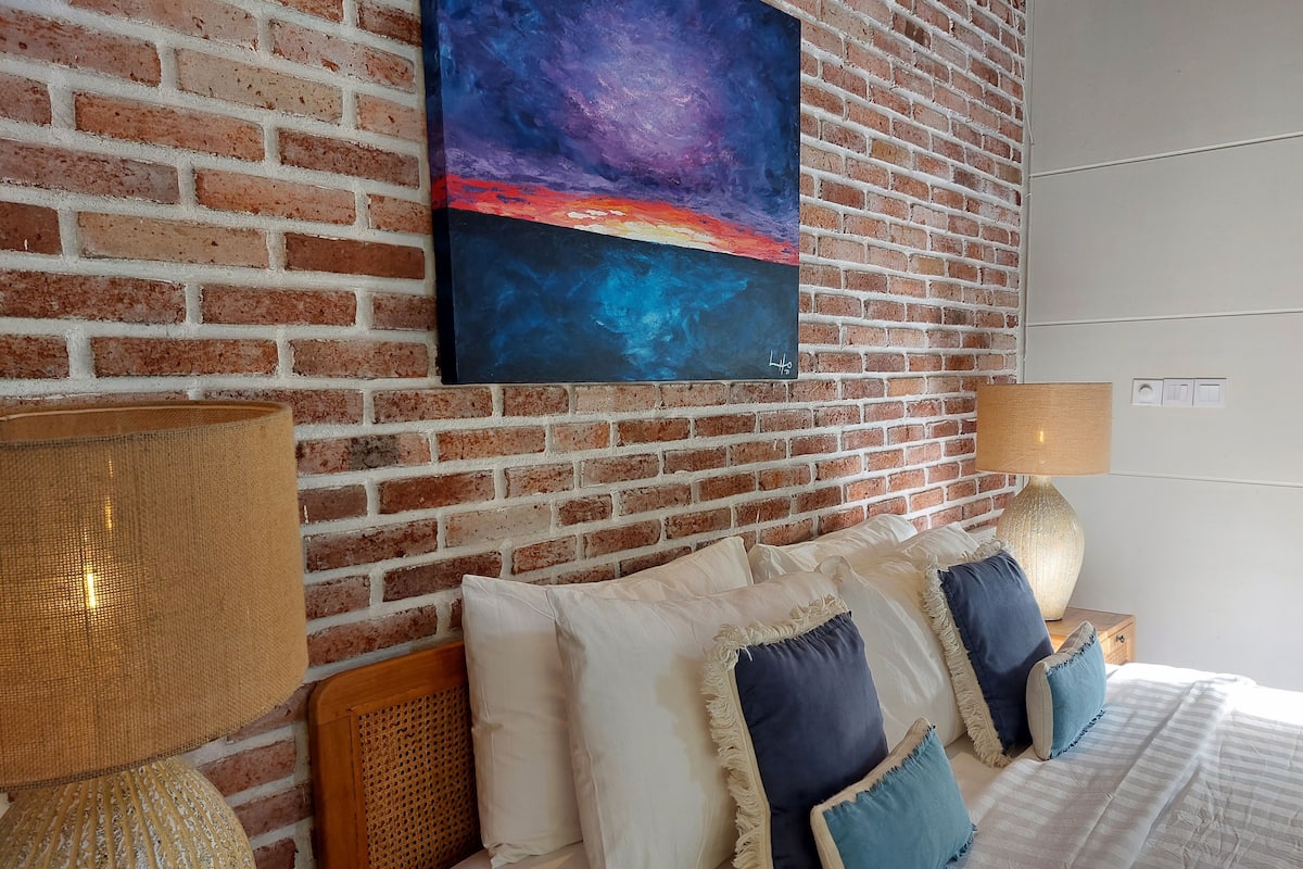A bedroom with a brick wall, a painting, a bed with multiple pillows, and a lamp.