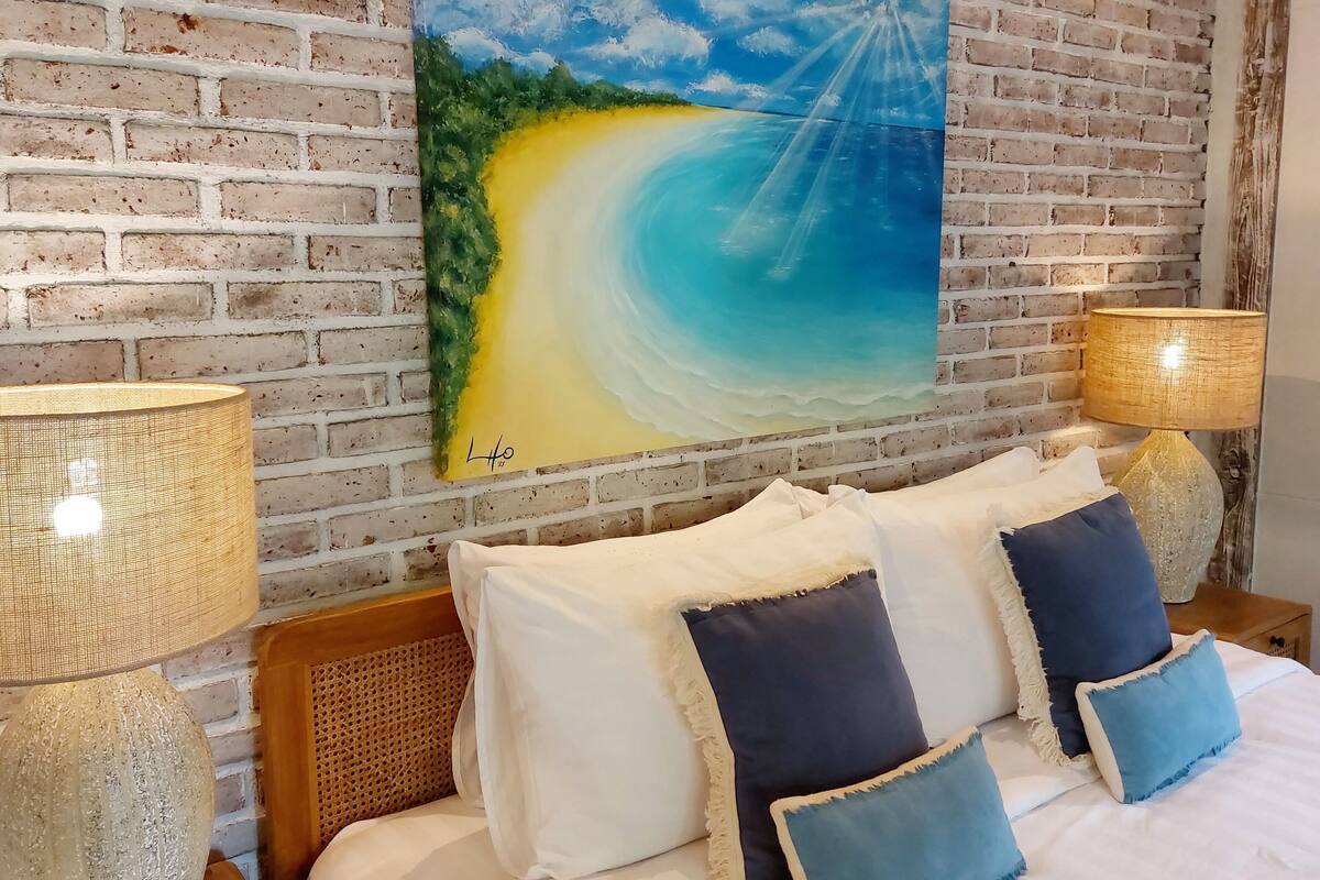 A bedroom with a brick wall, a painting of a beach, a bed with white and blue pillows, and two lamps on wooden nightstands.