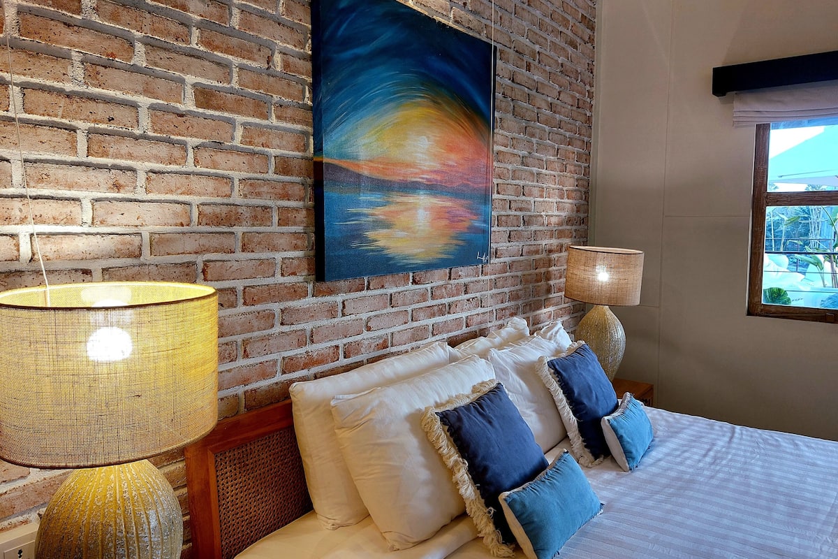 A hotel room with a brick wall, a painting, a bed with pillows, a lamp, and a window with a view.