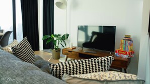 A modern living room with a flat-screen TV, a wooden side table with a remote, a lamp, and a stack of books. - New Year eve’s Fireworks & BayView Apt in CBD (Melbourne)