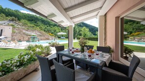 Um alpendre coberto com uma mesa posta para refeições, rodeado por cadeiras de vime, com vista para uma piscina e vegetação exuberante.