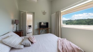 Ein Schlafzimmer mit einem Bett, einem Fernseher auf einem Schrank, einem Fenster mit Blick auf Grünflächen und einer Tür zu einem weiteren Raum.