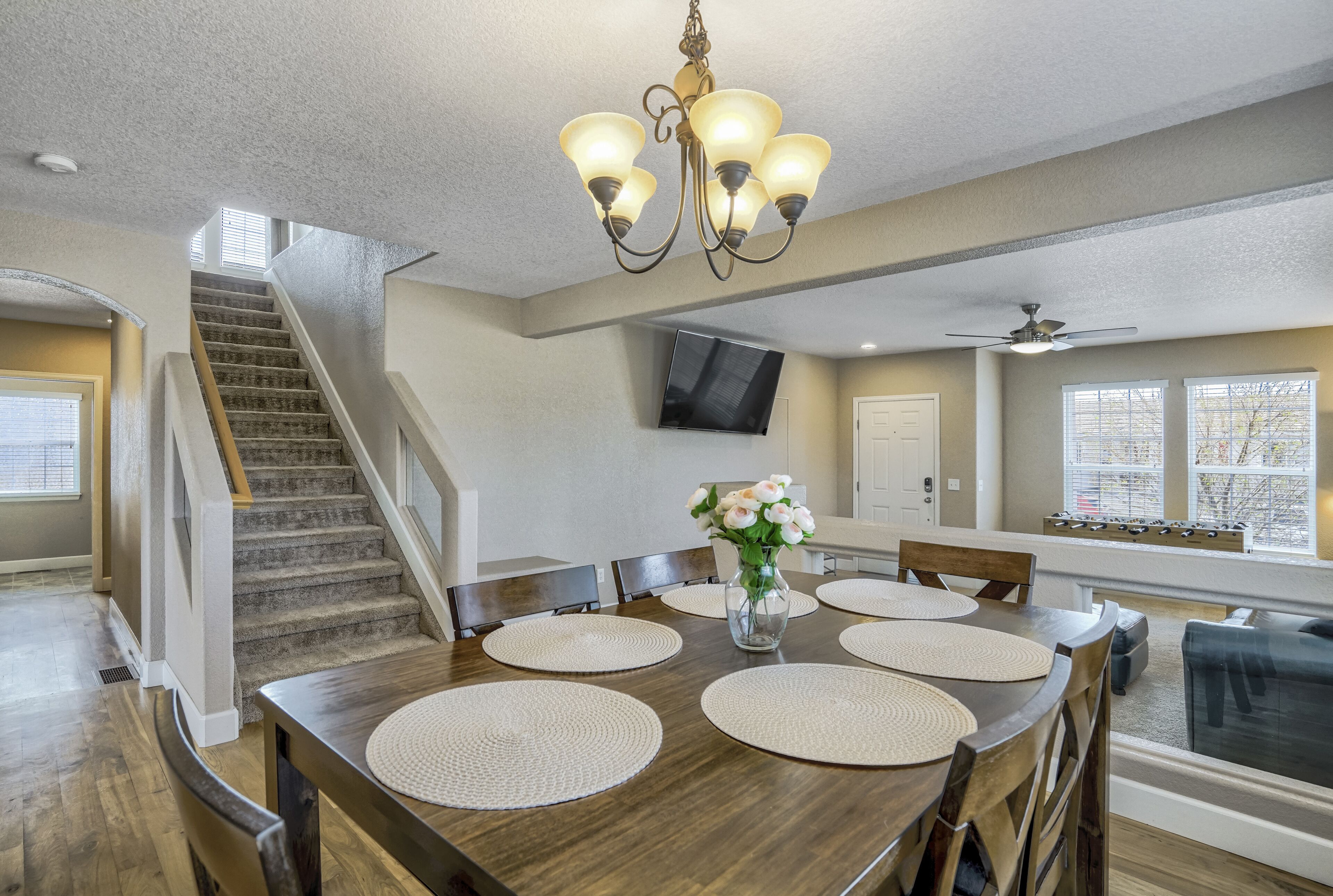 A dining area with a wooden table, woven placemats, and a vase of flowers. A staircase and a ceiling fan are visible in the background.