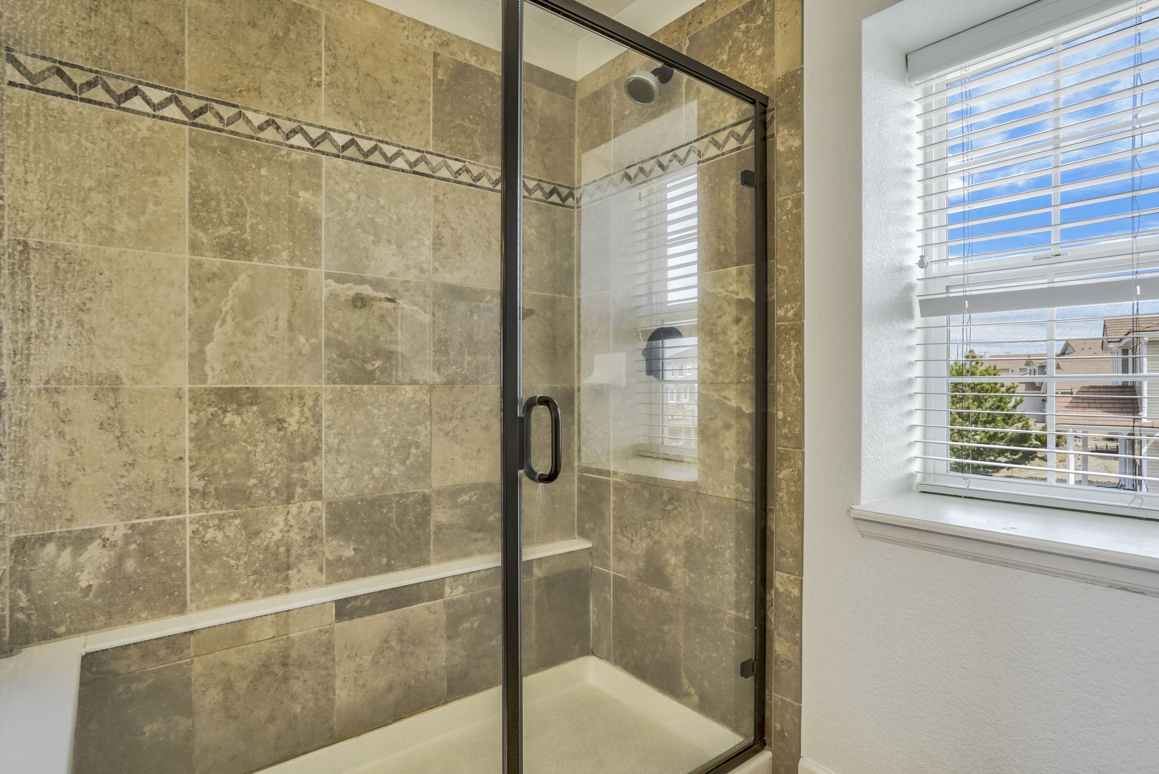 A glass-enclosed shower with beige tiles and a window with blinds.