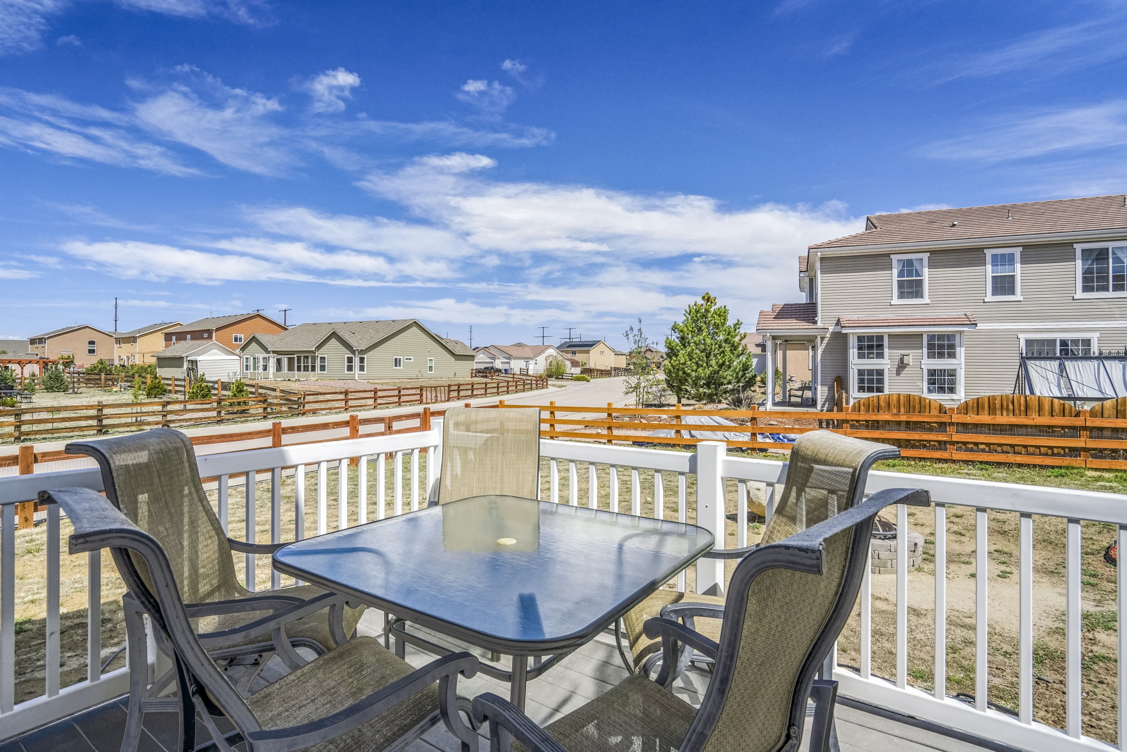 Una terraza con una mesa y sillas de vidrio, con vista a una zona residencial con casas y una valla de madera.
