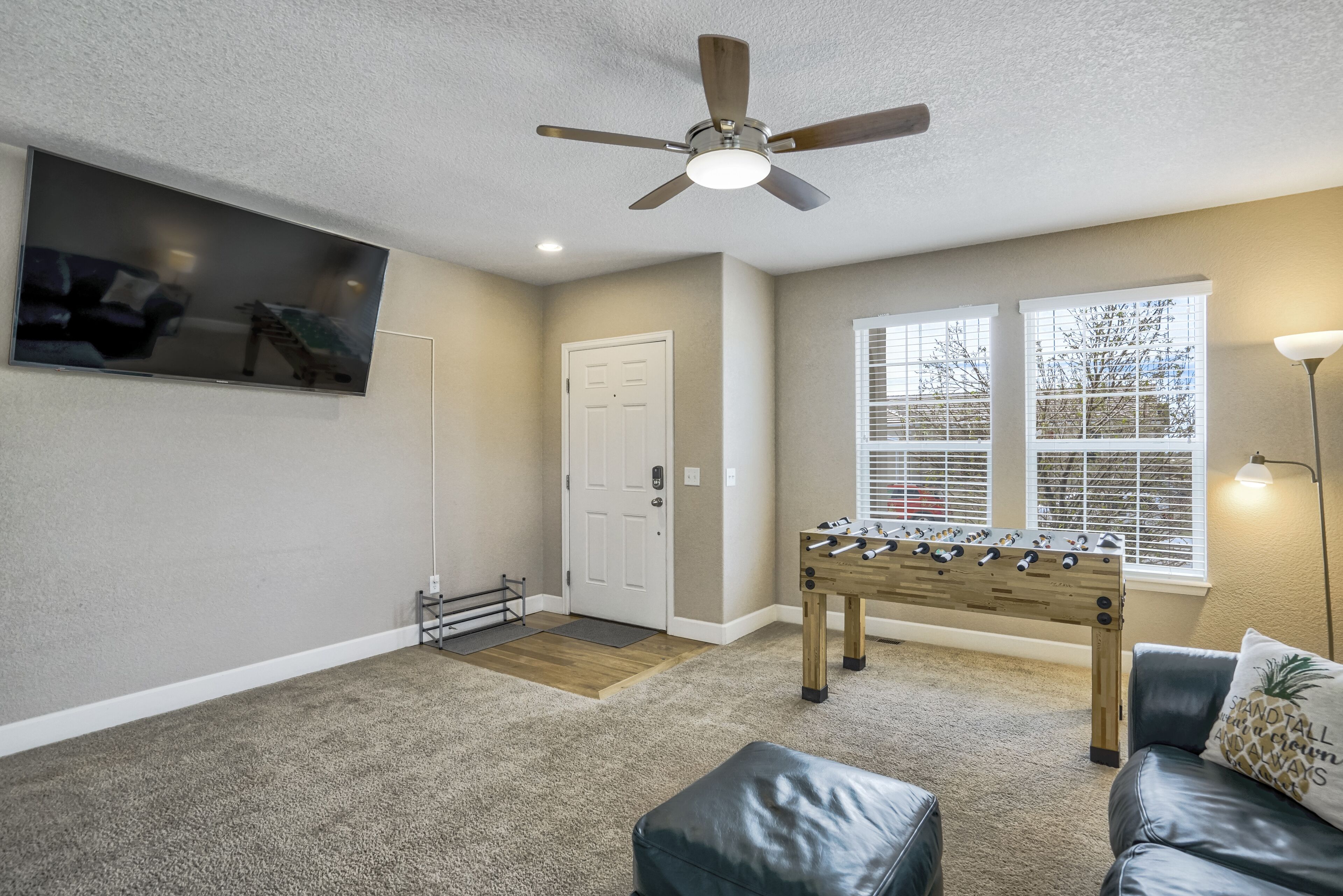 A living room with a foosball table, a ceiling fan, a TV mounted on the wall, and a window with blinds.