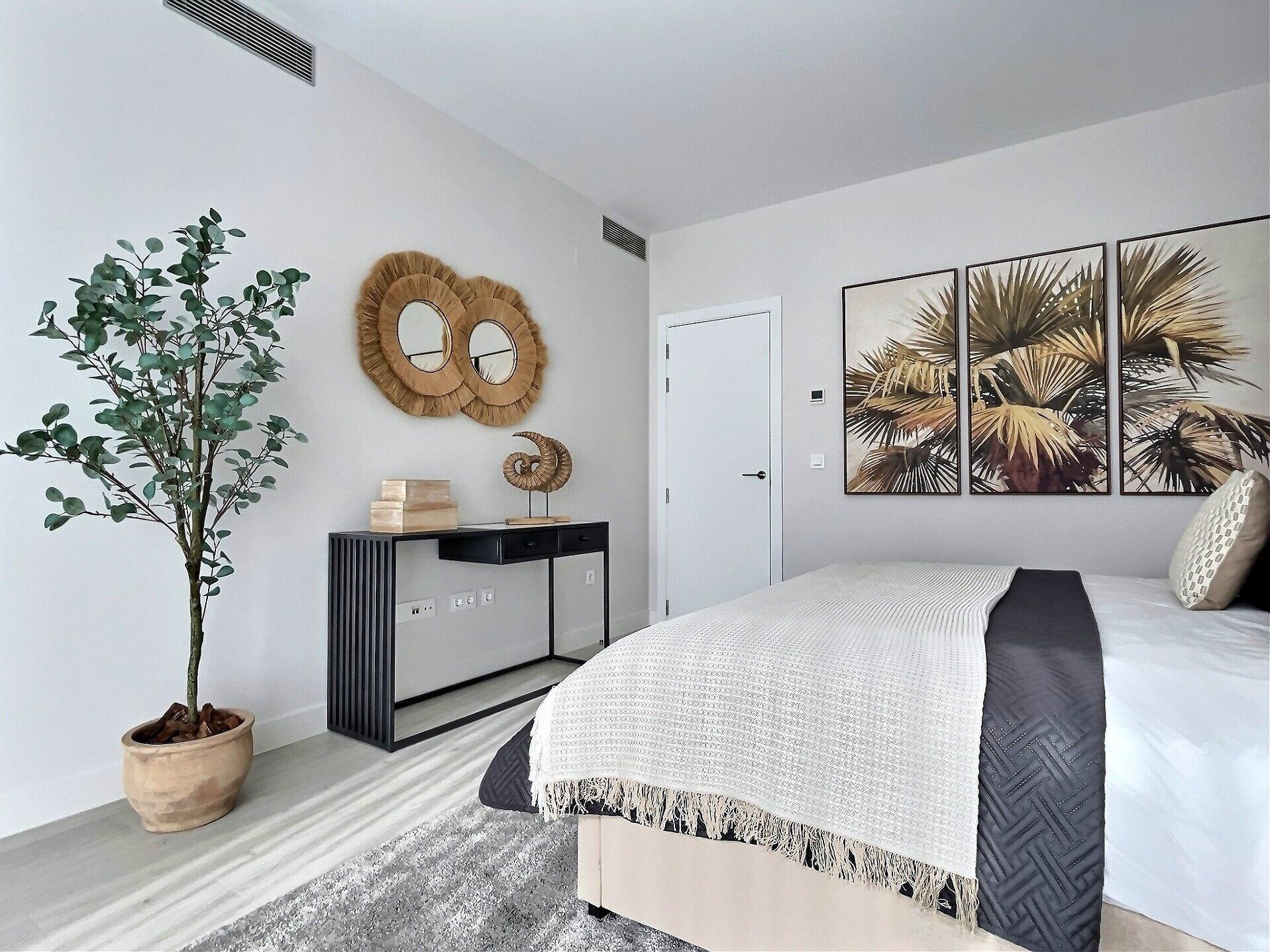 Un dormitorio moderno con una cama, una planta en maceta, una mesita de consola con espejos decorativos y una pared con pinturas de hojas tropicales.
