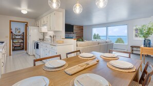 Una cocina moderna con armarios blancos, una mesa de comedor de madera dispuesta con platos, y vista al océano a través de ventanas amplias.