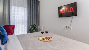 A hotel room with a bed, a TV mounted on the wall displaying 'NETFLIX', two wine glasses on the nightstand, and a plant in the corner. - Lumina apart near Michał Klepacz Park (Lodz)