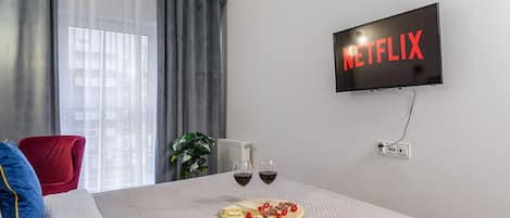 Habitación de hotel con una cama, un televisor montado en la pared mostrando 'NETFLIX', dos copas de vino sobre la mesita de noche y una planta en la esquina.