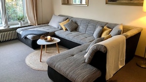 A modern living room with a grey sectional sofa, a small white side table, a floor lamp, and a painting on the wall. - 2 bedroom bungalow with kitchen, dining and living room with garden & terrace  (Bispingen)
