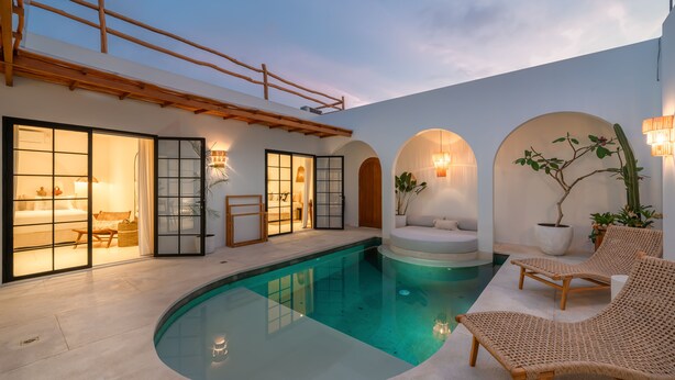 A modern indoor pool area with a curved pool, wooden lounge chairs, and a seating area with cushions. - Villa Valeria (Canggu)