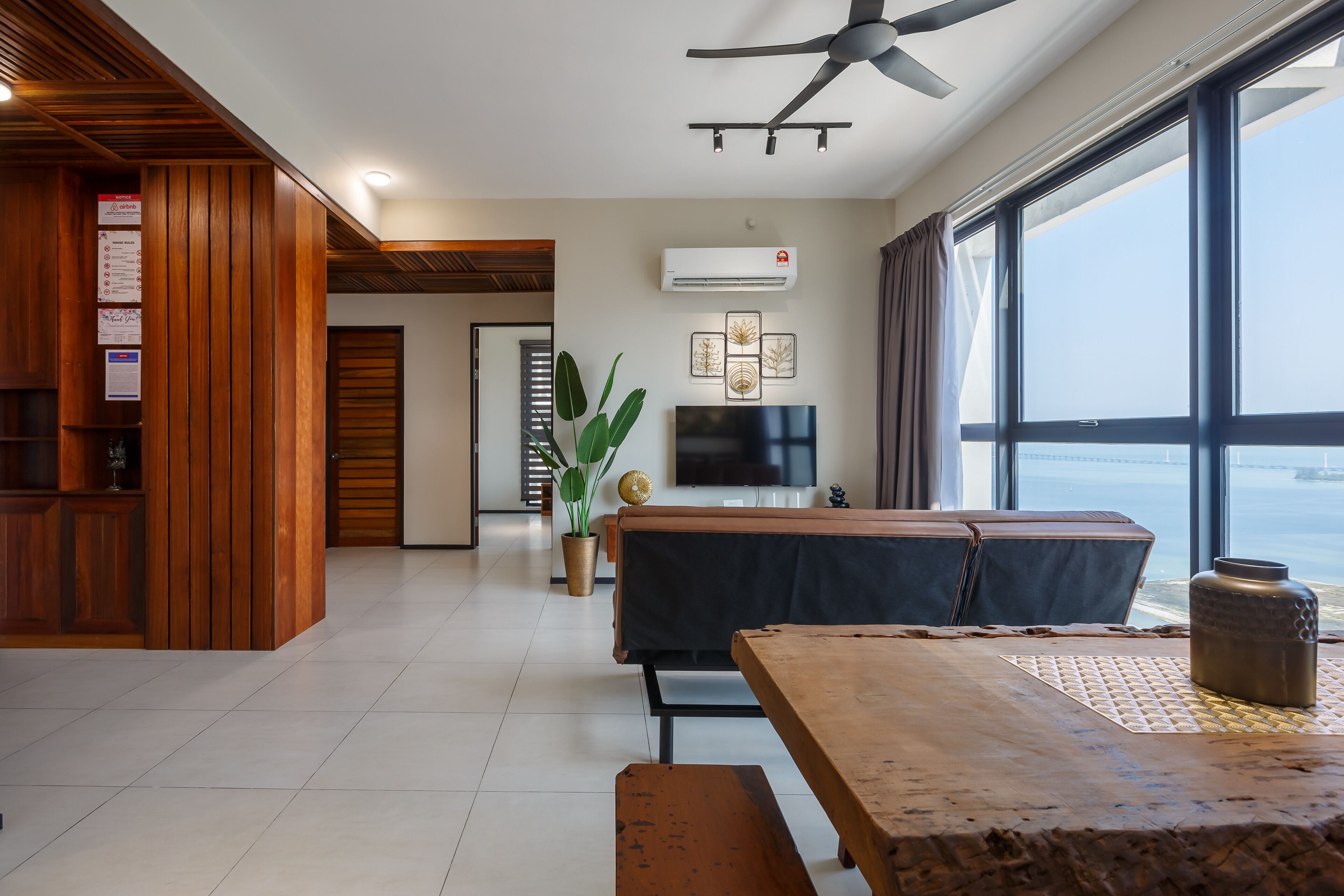 A modern living room with a wooden accent wall, a flat-screen TV, a large window with a view of the sea, and a ceiling fan.