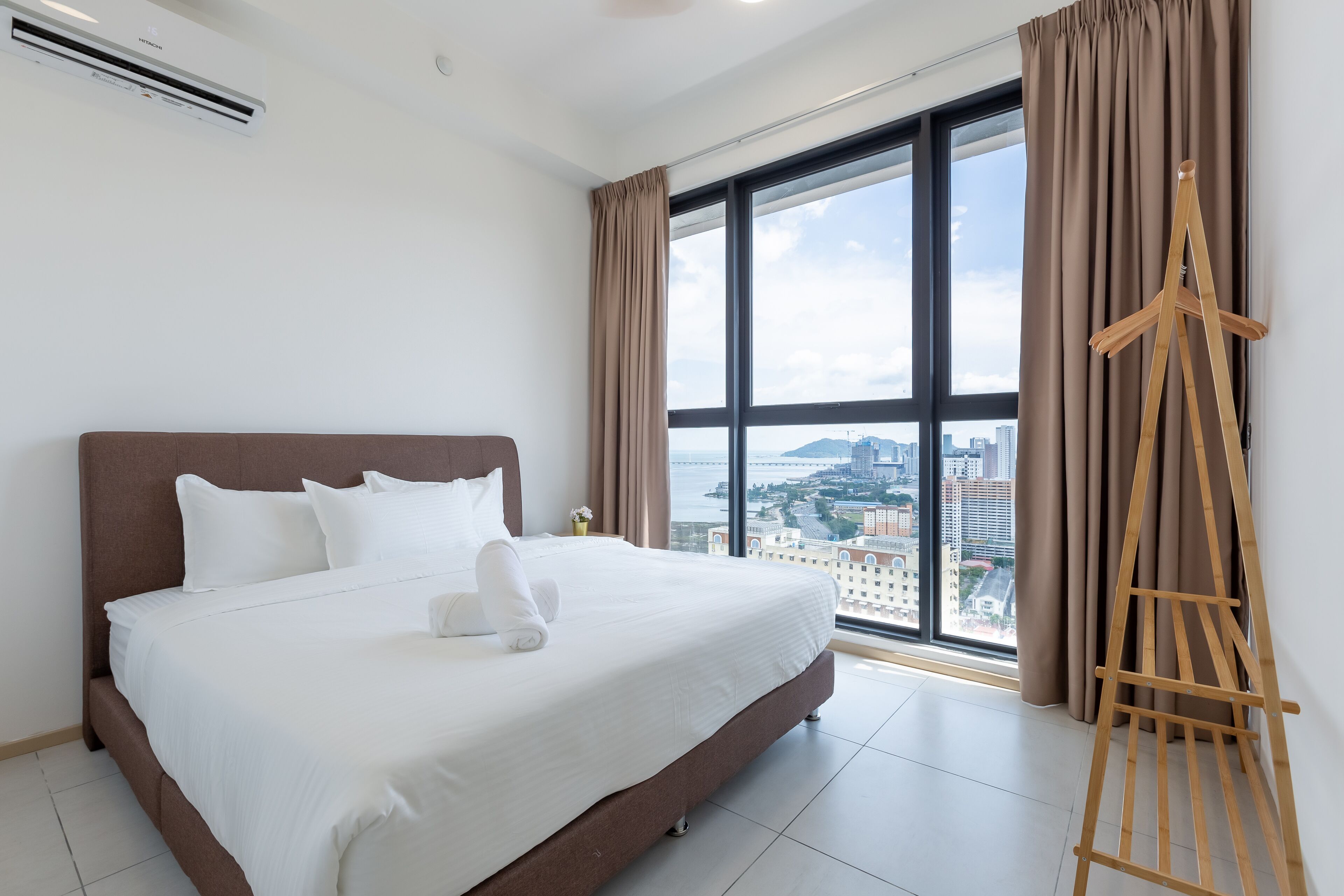 A hotel room with a large bed, a wooden easel, and a city view through large windows.