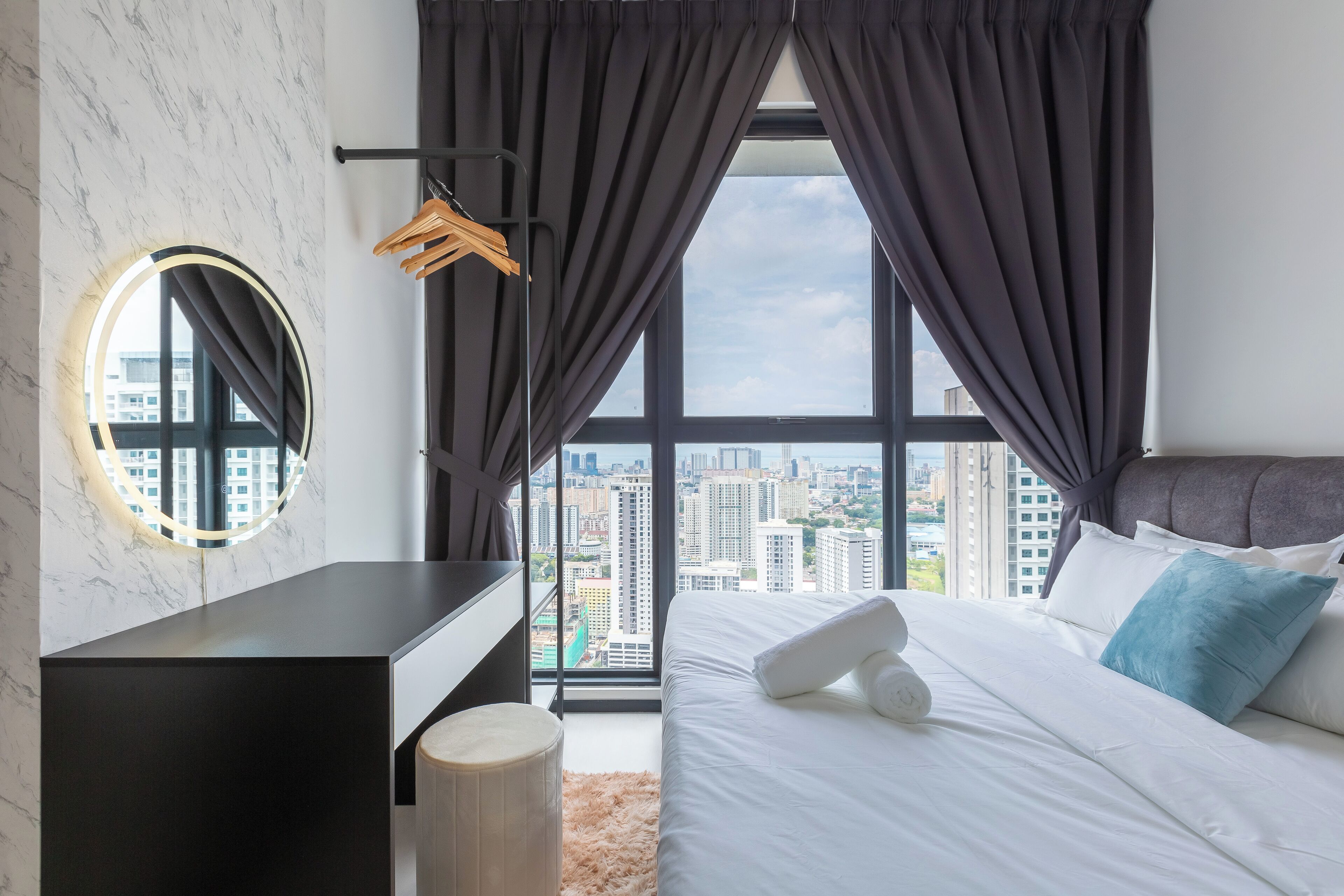 Quarto moderno com vista para a cidade, espelho redondo, mesa de cabeceira e cama com roupa de cama branca e almofada azul.