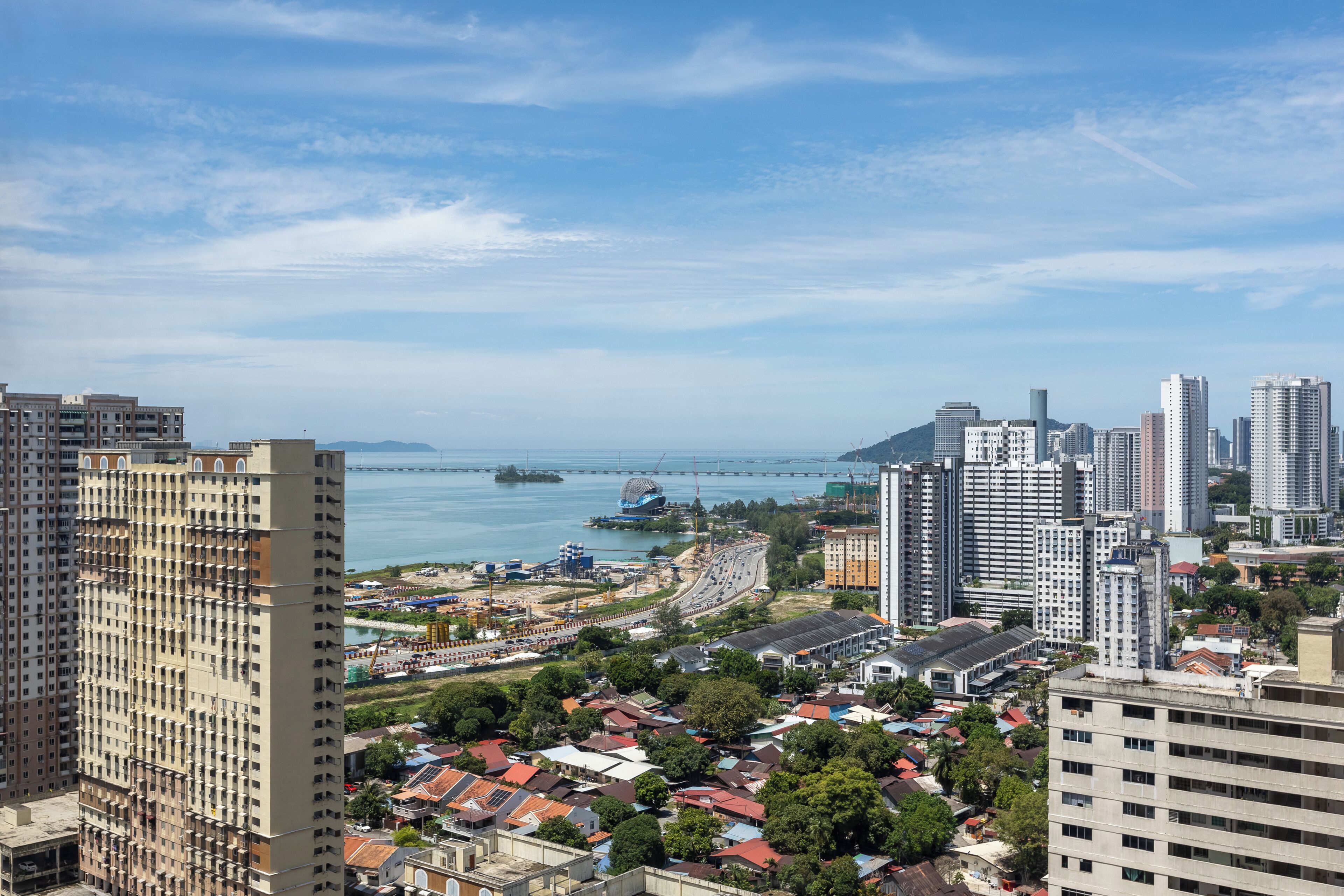 A high-rise building with multiple floors and balconies, surrounded by other high-rise buildings and a body of water with a bridge in the distance.