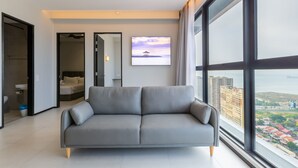 Una sala de estar moderna con un sofá gris, un ventanal con vistas a la ciudad y un cuadro de una playa.