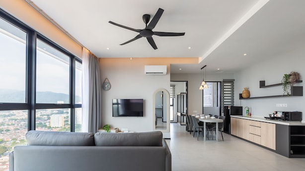Una sala de estar moderna con un sofá gris, un ventilador de techo y un amplio ventanal con vistas a la ciudad.
