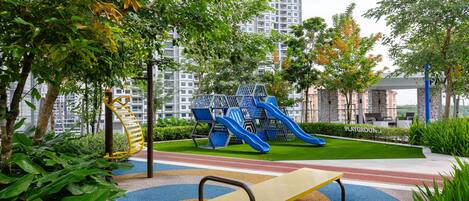 A playground with slides and a bench, surrounded by greenery and buildings in the background.