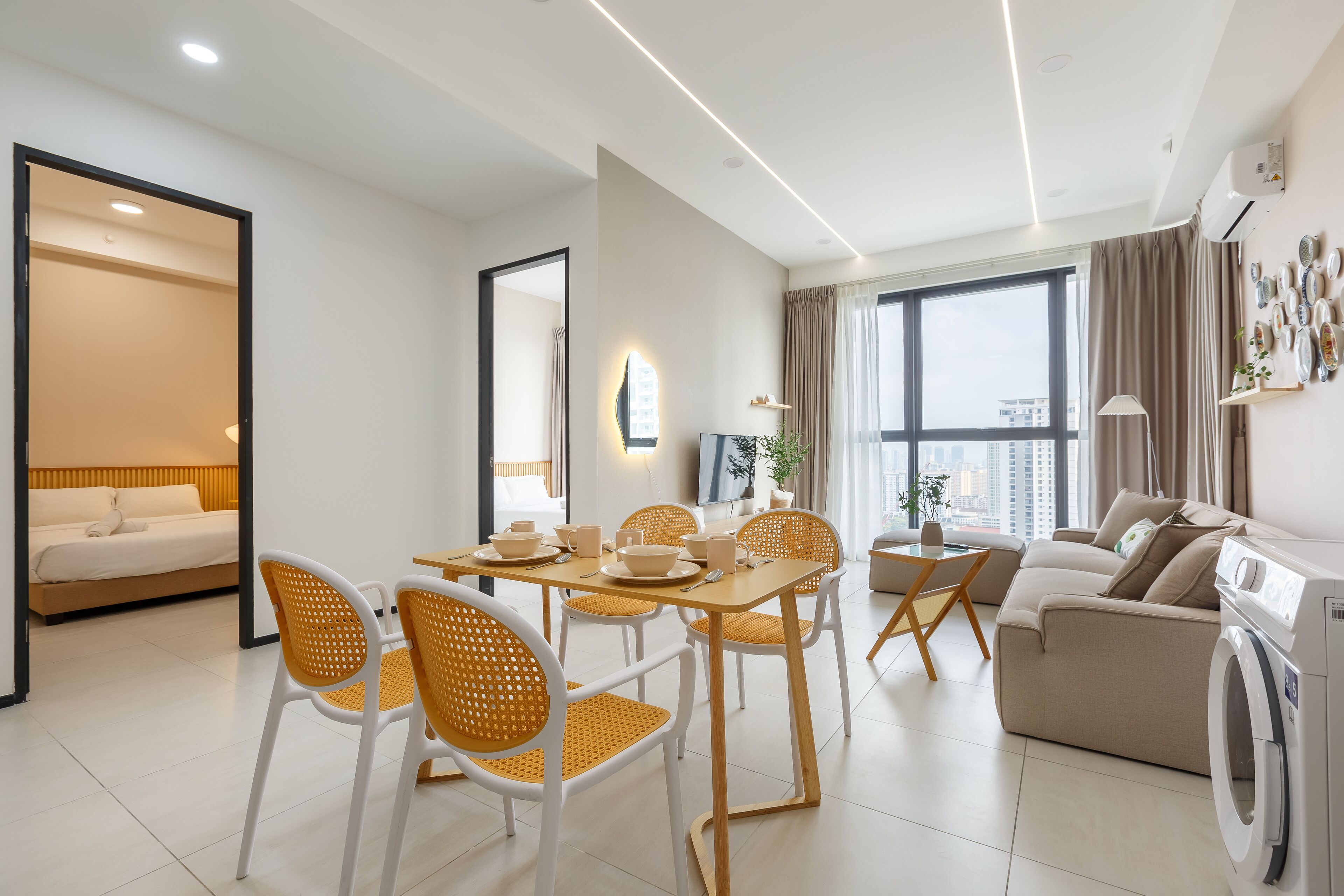 A modern, well-lit living room with a dining area, a sofa, a washing machine, and a large window offering a city view.